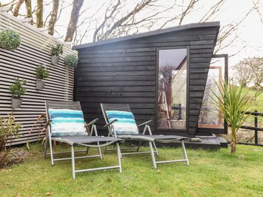 Two lounge chairs with pillows on grass near a black wooden shed and white slatted fence at Inny Brook Cottage in Davidstow near Camelford