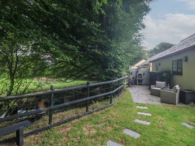 An outdoor patio area with two chairs and a table next to a building and a fenced grassy area with trees at Inny Brook Cottage Davidstow near Camelford