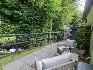 An outdoor patio with wicker armchairs a small table with wine and glasses a grill and a small metal table with chairs surrounded by trees at Inny Brook Cottage in Davidstow near Camelford