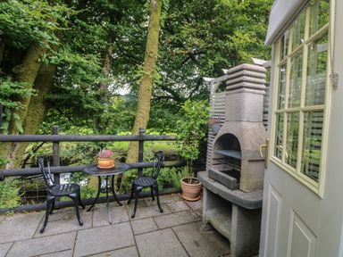 A patio with a small table and two chairs next to a stone barbecue and plants at Inny Brook Cottage in Davidstow near Camelford