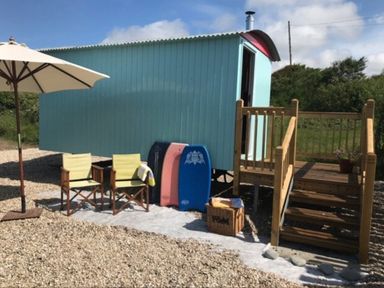 An outdoor area with a light blue wooden hut on stilts with stairs and railing two chairs under a large white umbrella and three bodyboards leaning against the hut at Shepherd's Hut in Southerndown near St Brides Major