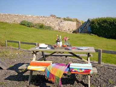 An outdoor picnic table with bottles glasses and folded blankets on benches in a garden with a stone wall and small building in the background at Shepherd's Hut in Southerndown near St Brides Major
