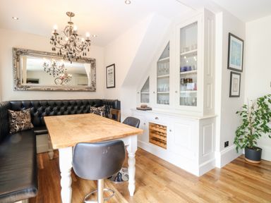 A dining area with a wooden table black leather bench seating chandelier and white cabinet with glass doors at The Farmhouse in Felmingham near North Walsham