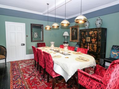 A dining room with a long table covered with a white cloth surrounded by red patterned chairs and a red rug at The Farmhouse in Felmingham near North Walsham
