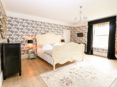 A bedroom with a large cream bed black lamps a chandelier and patterned wallpaper at The Farmhouse in Felmingham near North Walsham