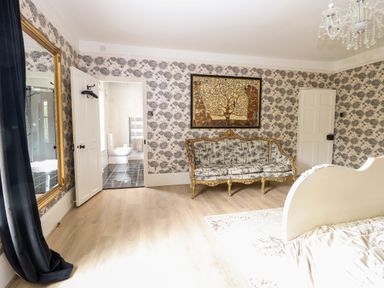 A bedroom with patterned wallpaper a decorative sofa a large mirror a chandelier and an open door leading to a bathroom at The Farmhouse in Felmingham near North Walsham