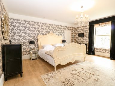 A bedroom with a cream bed frame a chandelier patterned wallpaper and a window with black curtains at The Farmhouse in Felmingham near North Walsham