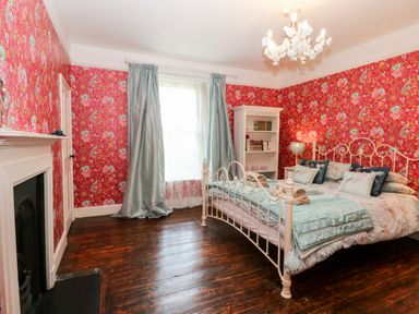 A bedroom with a white metal bed framed by floral bedding a bookshelf curtains and a fireplace at The Farmhouse in Felmingham near North Walsham