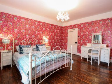 A bedroom with a white metal bed frame pillows and a teal blanket bedside tables with lamps a white dressing table and chair on wooden floors with floral wallpaper at The Farmhouse in Felmingham near North Walsham