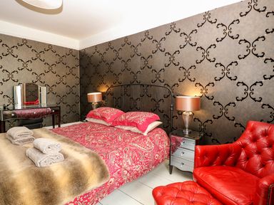 A bedroom with a bed covered in red patterned bedding two bedside tables with lamps a red armchair and ottoman and a dressing table with mirror at The Farmhouse in Felmingham near North Walsham
