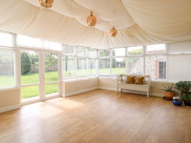 A sunroom with wooden flooring a white bench with cushions potted plants and large windows with blinds at The Farmhouse Felmingham near North Walsham