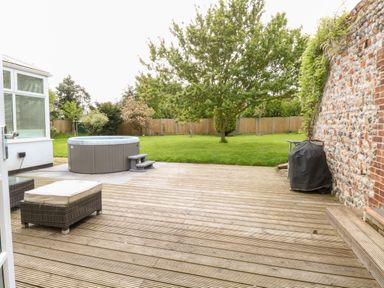 A wooden deck with a hot tub ottoman and covered barbecue next to a grass lawn with trees at The Farmhouse in Felmingham near North Walsham