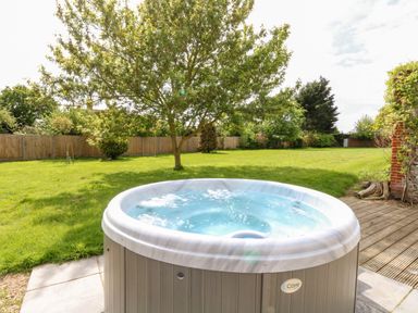 An outdoor hot tub on a patio next to a lawn with a tree and wooden fence at The Farmhouse in Felmingham near North Walsham