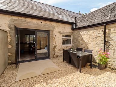 An outdoor patio area with a glass table and six chairs on gravel outside a stone house at Mayfield Cottage Nr Malmesbury