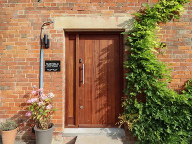 A wooden front door on a brick wall with green ivy and potted plants at Mayfield Cottage Nr Malmesbury