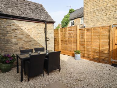 An outdoor patio area with a glass table and four black chairs on a gravel floor at Mayfield Cottage Nr Malmesbury
