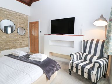 A bedroom with a bed, towels, a striped armchair, a television mounted on a white wall, two round mirrors on a stone wall, and a wooden door at Mayfield Cottage Nr Malmesbury