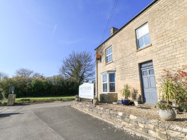 A stone building with a grey door and bay window next to a sign on a roadside at Mayfield Cottage in Nr Malmesbury