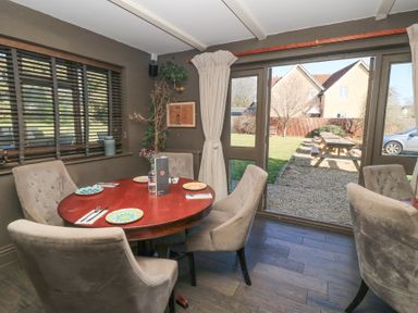 A dining area with a round wooden table set with plates and cutlery and four upholstered chairs next to large windows and glass doors opening to an outdoor area with picnic tables at Mayfield Cottage in Nr Malmesbury