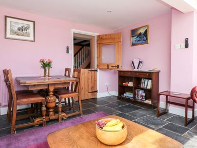 A dining area with a wooden table and chairs a wooden bowl with fruit on a coffee table and a wooden cabinet with books on shelves at The Old Forge Cottage in Kilkhampton