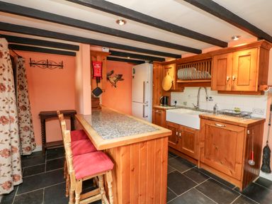 A kitchen with wooden cabinets a granite countertop a white farmhouse sink and two wooden stools with red cushions at The Old Forge Cottage in Kilkhampton