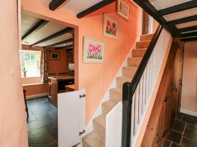 A staircase with carpeted steps and wooden railing next to wooden storage cabinets and an open door leading to a room with a window and chairs at The Old Forge Cottage in Kilkhampton