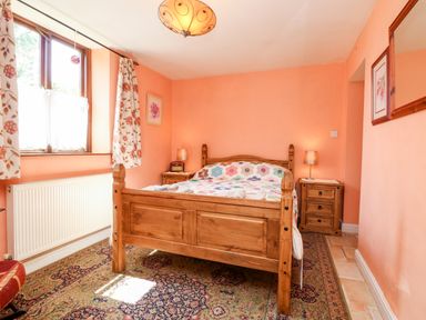 A bedroom with a wooden bed two bedside tables lamps window with floral curtains and a patterned rug at The Old Forge Cottage in Kilkhampton