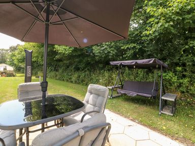 A garden patio with a table and chairs under an umbrella and a swing seat nearby at The Old Forge Cottage in Kilkhampton