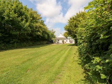 A large grassy yard with trees on both sides and a small house with a patio at The Old Forge Cottage in Kilkhampton