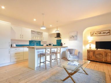 A kitchen area with white cabinets, a wooden countertop island with two wooden stools, a chair, a glass coffee table, and a wooden TV stand with a lamp and television at The Loft Amble