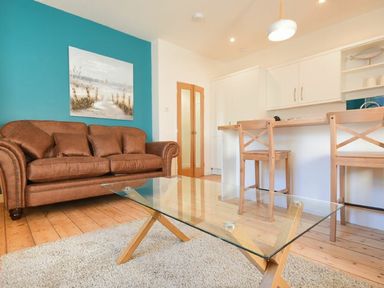 A living room with a brown sofa a glass coffee table and a kitchen counter with wooden stools at The Loft (Amble) in Amble