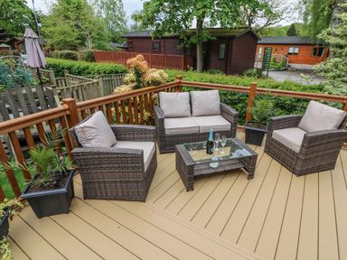An outdoor wooden deck with rattan sofa and chairs around a glass coffee table with a bottle and glasses at 8 Waterside Wood White Cross Bay near Troutbeck Bridge