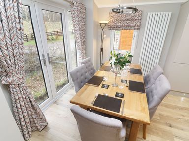 A dining area with a wooden table set for six and gray upholstered chairs by glass doors at 8 Waterside Wood White Cross Bay near Troutbeck Bridge