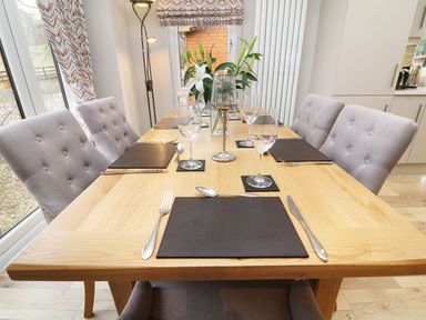 A dining room with a wooden table set with gray placemats glassware and cutlery surrounded by gray cushioned chairs with a window and curtains in the background at 8 Waterside Wood White Cross Bay near Troutbeck Bridge