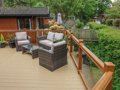 An outdoor wooden deck with wicker chairs and a glass table next to wooden cabins and greenery at 8 Waterside Wood White Cross Bay near Troutbeck Bridge