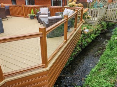 An outdoor wooden deck with glass railing overlooking a small stream with greenery surrounding it at 8 Waterside Wood White Cross Bay near Troutbeck Bridge
