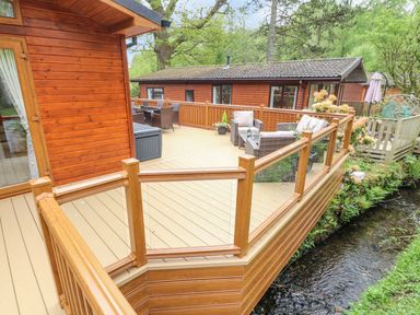 A wooden deck with outdoor furniture and glass railing next to a small stream at 8 Waterside Wood in White Cross Bay near Troutbeck Bridge