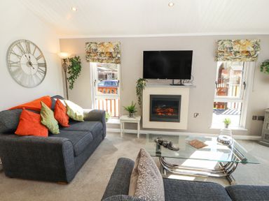 A living room with a grey sofa with orange and green cushions a glass coffee table a wall-mounted TV and an electric fireplace at Bowness 66 in Troutbeck Bridge