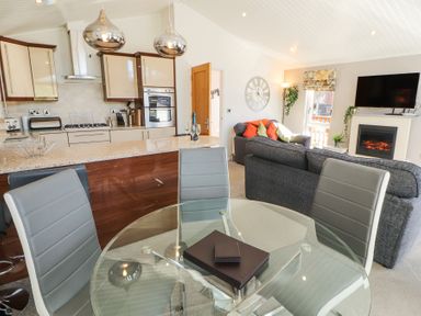 An open-plan living area with a kitchen, glass dining table, chairs, sofas, fireplace, and television at Bowness 66 in Troutbeck Bridge