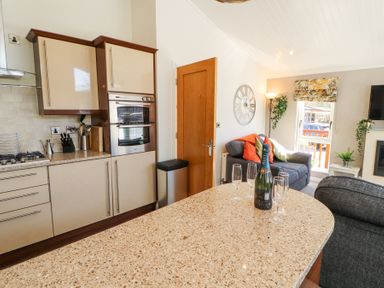 A kitchen with a countertop holding four wine glasses and a bottle of champagne next to a living area with a grey sofa and a large clock on the wall at Bowness 66 in Troutbeck Bridge