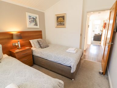 A bedroom with two single beds a wooden nightstand with a lamp paintings on the wall and an open door showing a living area at Bowness 66 in Troutbeck Bridge