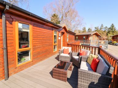 An outdoor deck with wicker seating and cushions outside wooden lodges at Bowness 66 in Troutbeck Bridge
