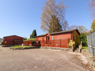 A wooden cabin with a deck and railing surrounded by a gravel driveway and trees at Bowness 66 in Troutbeck Bridge