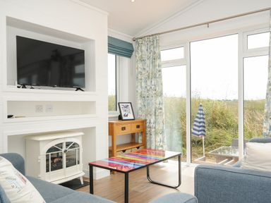 A living room with a television above a white electric heater two blue sofas and a colorful coffee table by a wooden side table with a framed picture and large windows with floral curtains at Midway Lodge in Cawsand near Kingsand