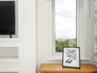 A window with a view of greenery and a body of water beside a wooden cabinet with a framed artwork a TV on a white wall in Midway Lodge in Cawsand near Kingsand