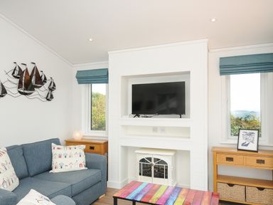 A living room with a blue couch a colorful coffee table a wall-mounted TV and wooden side tables at Midway Lodge in Cawsand near Kingsand