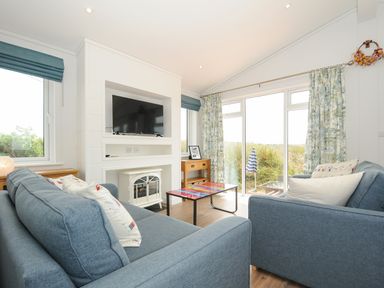 A living room with two blue sofas a TV on a white built-in unit and glass doors with floral curtains at Midway Lodge in Cawsand near Kingsand