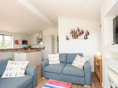 A living room with blue sofas and cushions next to a white kitchen at Midway Lodge in Cawsand near Kingsand