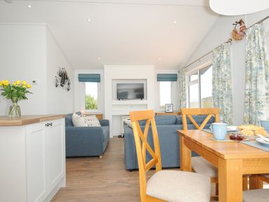 A living room with blue sofas wooden dining table and chairs a TV and large windows with curtains at Midway Lodge in Cawsand near Kingsand