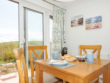 A dining area with a wooden table and chairs set with plates coffee mugs and croissants by a glass sliding door with curtains at Midway Lodge in Cawsand near Kingsand